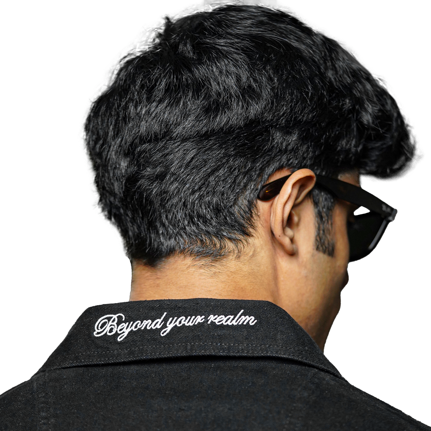 A close-up of the back of a black denim jacket with the text "Beyond your realm" embroidered on the collar. The person wearing the jacket has short, dark hair and is wearing sunglasses.