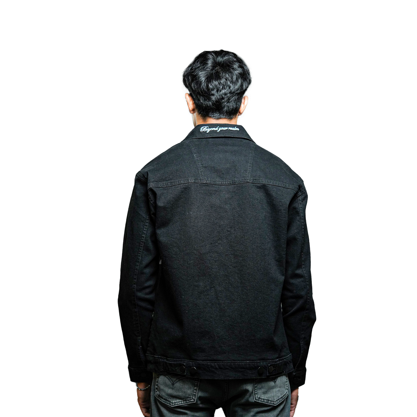 A full view of the back of a person wearing a black denim jacket with the embroidered text "Beyond your realm" on the collar. The jacket is paired with dark pants, and the person stands with their back to the camera.