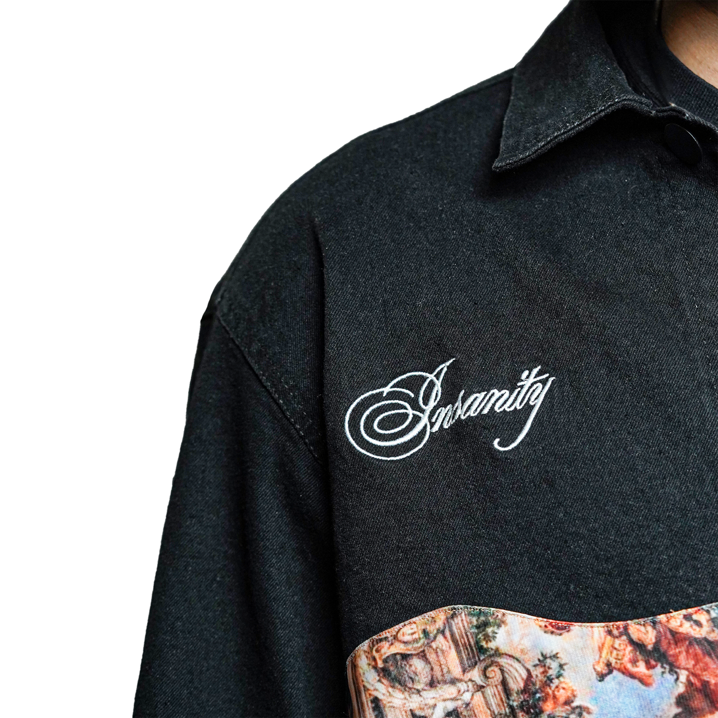 A close-up shot of a black denim jacket showcasing white embroidered text "Insanity" on the chest and a printed artistic design on the front panel.