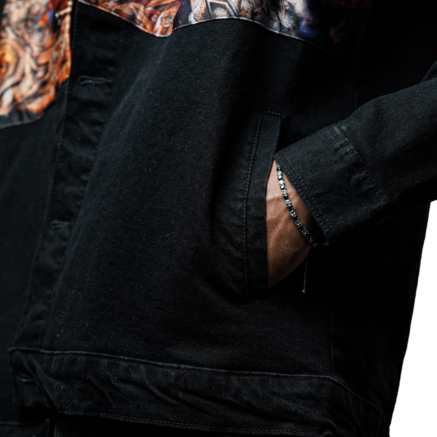A close-up of a person wearing a black denim jacket with the hand placed in the pocket. A silver bracelet with small stones is visible on the wrist.