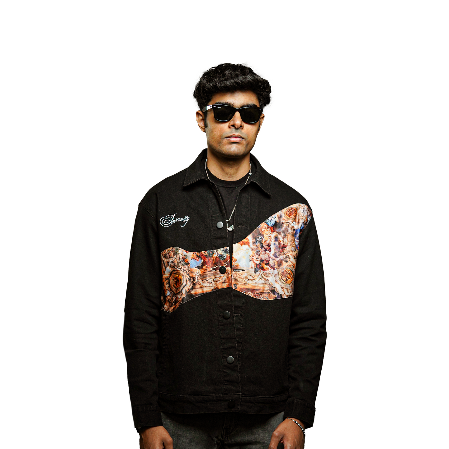 A man with dark hair and sunglasses is wearing a black denim jacket with an artistic printed design across the chest and upper sleeves. The design features a classical painting-style motif with warm tones, contrasting against the solid black fabric. The word "Serenity" is embroidered in white on the left side of the chest. He has a confident expression, accessorized with a silver chain necklace and a bracelet. The background is plain white.