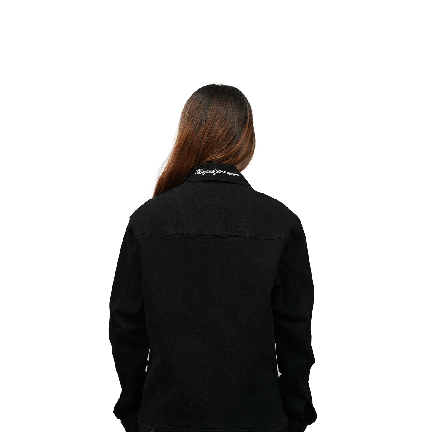 A female model standing with her back to the camera, wearing a black denim jacket with white embroidered text "Beyond your realm" on the collar. The jacket is plain black on the back.
