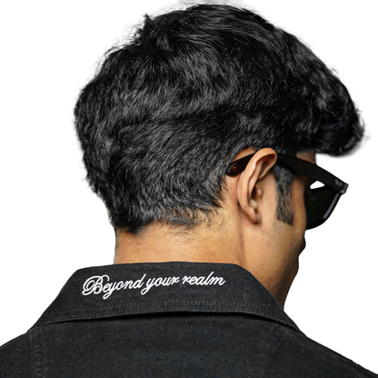 A close-up of the back of a black denim jacket with the text "Beyond your realm" embroidered on the collar. The person wearing the jacket has short, dark hair and is wearing sunglasses.