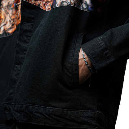 A close-up of a person wearing a black denim jacket with the hand placed in the pocket. A silver bracelet with small stones is visible on the wrist.